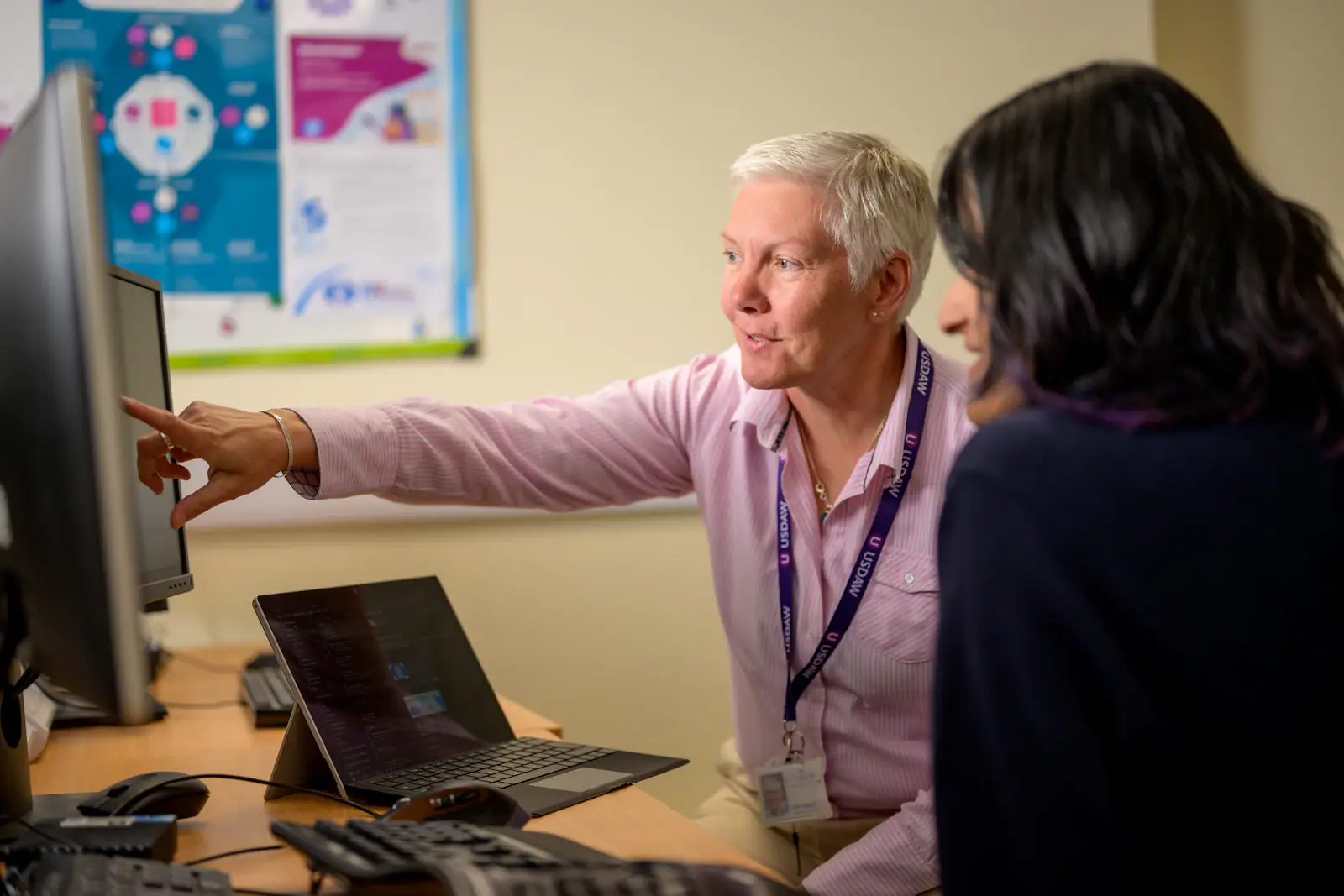 A colleague points at a laptop screen while explaining something to a teammate.